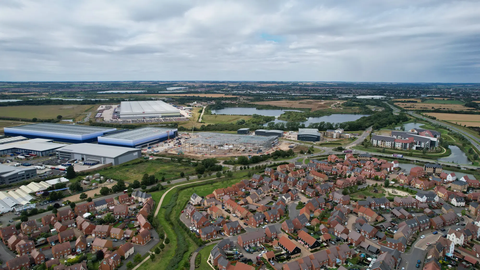 Warehouses with wide flat roofs beside a new estate of brick houses on the edge of a town, seen from the air on an overcast day