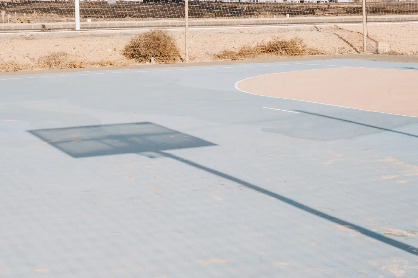 An empty paved court under full sun, casting a long hard shadow across the bare surface.